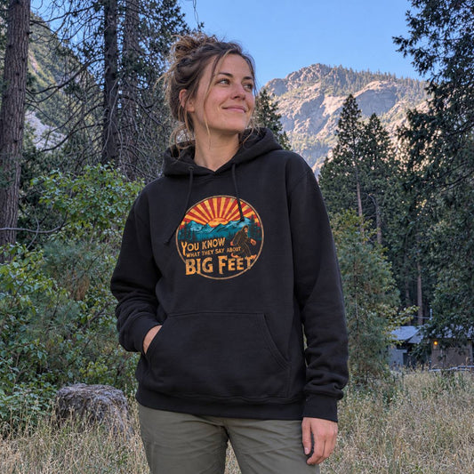 A woman wearing a black pullover hoodie with a retro mountain graphic and the text 'YOU KNOW WHAT THEY SAY ABOUT BIG FEET' stands outdoors in a forest with mountains in the background.