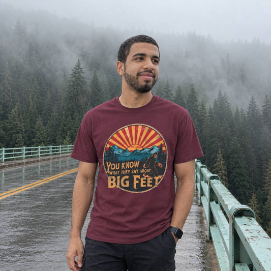 A man wearing a maroon graphic t-shirt with a mountain and sun design that reads 'YOU KNOW WHAT THEY SAY ABOUT BIG FEET', standing on a wet road with a misty forest background.