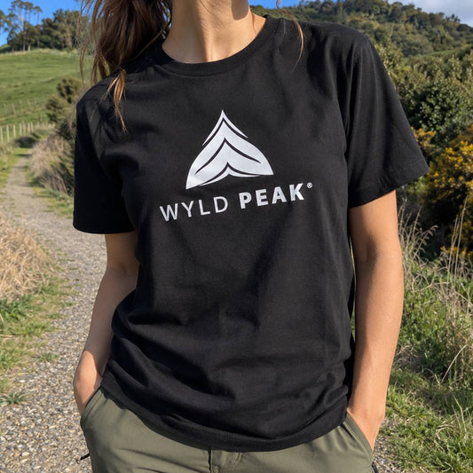 A person outdoors wearing a black short-sleeve T-shirt with a white WYLD PEAK logo and text on the chest, paired with olive green pants. The background shows a gravel path and grassy hills under bright daylight.