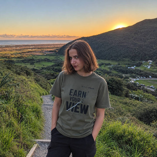 A woman stands outdoors on a hillside path at sunset wearing an olive green graphic T-shirt with the text 'EARN YOUR VIEW' and 'WYLD PEAK' printed on the front, paired with black pants. The background shows a scenic landscape with a valley, ocean, and mountains during golden hour.