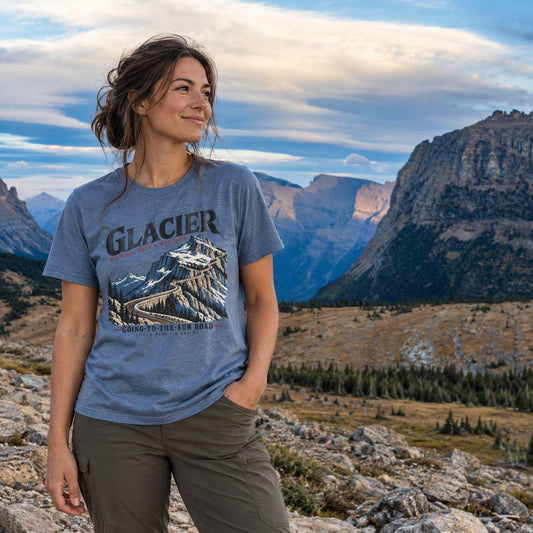 A woman wearing a slate blue graphic t-shirt with a mountain print and the word 'GLACIER' stands outdoors in a mountain landscape. The t-shirt features a graphic of a snow-capped mountain with the text 'GLACIER'.