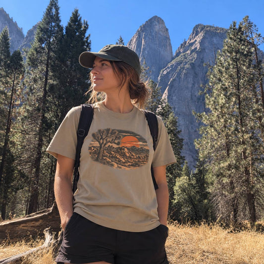 A woman wearing a beige graphic t-shirt, black shorts, and a dark green cap stands in a forest with tall mountains and a blue sky in the background.