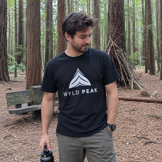 A man standing in a redwood forest wearing a black crewneck t-shirt with a white geometric mountain logo and the text 'WYLD PEAK' printed on the chest.