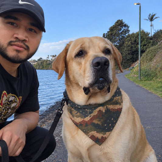 A golden retriever dog wearing a camouflage bandana sits on a paved path next to a man wearing a black Nike cap and t-shirt, with water and trees in the background.
