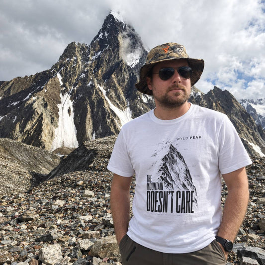 A man wearing a white graphic t-shirt with 'THE MOUNTAIN DOESN'T CARE' and 'WYLD PEAK' printed on the front, standing in a rocky mountain landscape with a sharp peak in the background.