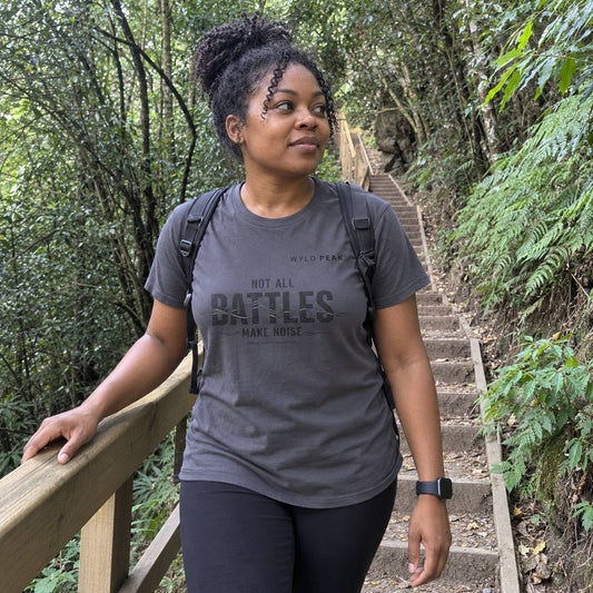 A woman wearing a dark gray graphic t-shirt with 'NOT ALL BATTLES MAKE NOISE' printed on the front, standing on a wooden railing on a stone staircase in a lush green forest.