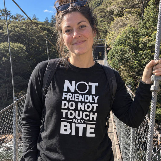 A woman wearing a black long-sleeve t-shirt with a bold white graphic that reads 'NOT FRIENDLY DO NOT TOUCH MAY BITE' stands on a suspension bridge outdoors with trees in the background.