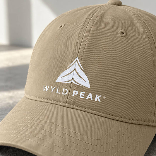 A close-up of a khaki baseball cap featuring a white embroidered WYLD PEAK logo on the front, set against a softly blurred light gray background.