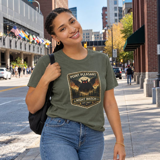 A woman walking on a city sidewalk wearing an olive green graphic t-shirt with a 'Point Pleasant Night Watch' design, paired with blue jeans and carrying a black shoulder bag.