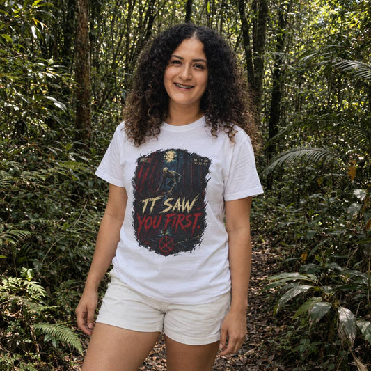 A woman with curly dark hair stands in a dense green forest wearing a white graphic t-shirt and light beige shorts. The t-shirt features a dark, distressed graphic with the text 'IT SAW YOU FIRST' in red and yellow.