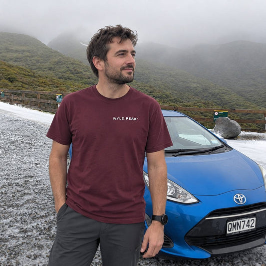 A man wearing a burgundy t-shirt with 'WYLD PEAK.' printed in white on the chest stands outdoors next to a blue Toyota car, with misty green mountains in the background.