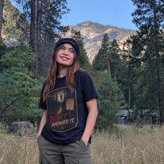 A woman wearing a black graphic t-shirt with the text 'DON'T ANSWER IT.' and a black beanie stands outdoors in a grassy field with a forest and mountain in the background.