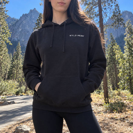 A woman wearing a black pullover hoodie with 'WYLD PEAK' printed in white on the chest, standing outdoors in a mountain forest setting.