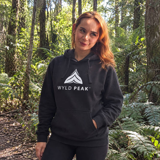 A woman with red hair stands in a lush green forest wearing a black pullover hoodie with a white 'WYLD PEAK' logo on the chest.