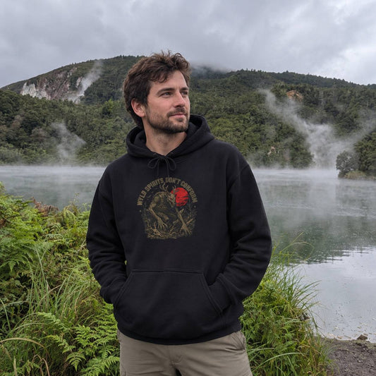 A man wearing a black pullover hoodie with a vintage-style graphic on the chest stands in front of a misty lake and forested mountains. The hoodie graphic features a circular design with a red sun, a skinwalker and the text 'WYLD SPIRITS DON'T CONFORM'.