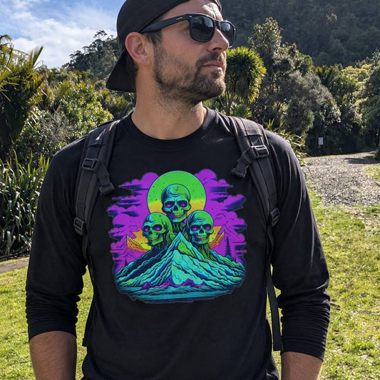 A man wearing a black long-sleeve t-shirt with a colorful graphic of three skulls on a mountain under a green sun, photographed outdoors in a grassy park.
