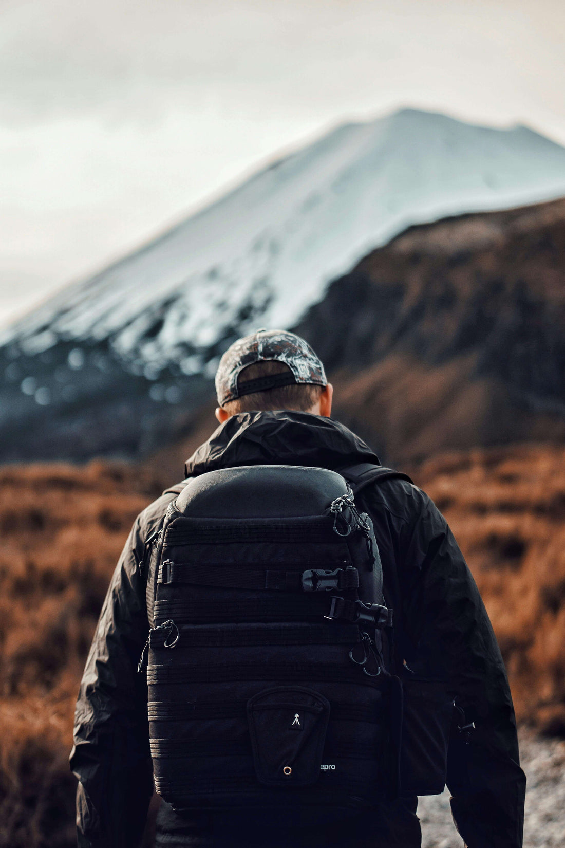 Hiker in cold weather gear with a tactical backpack standing before a snow-capped volcano on a rugged trail
