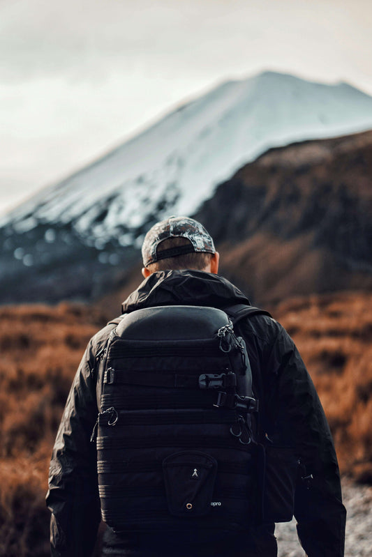 Hiker in cold weather gear with a tactical backpack standing before a snow-capped volcano on a rugged trail