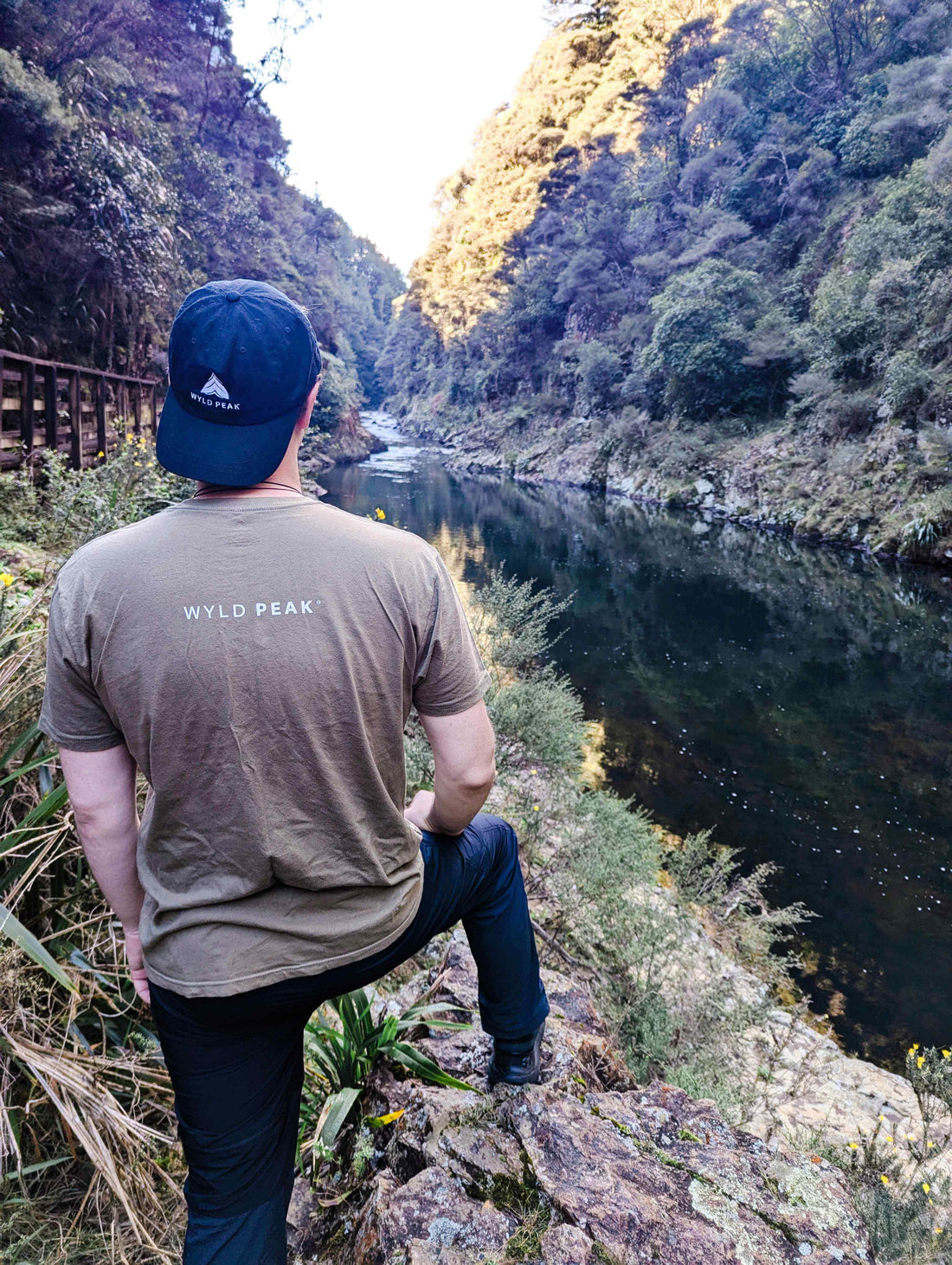 Alexander standing in a Wyld Peak tee, gazing out over the river at Karangahake Gorge, New Zealand, surrounded by rugged natural beauty