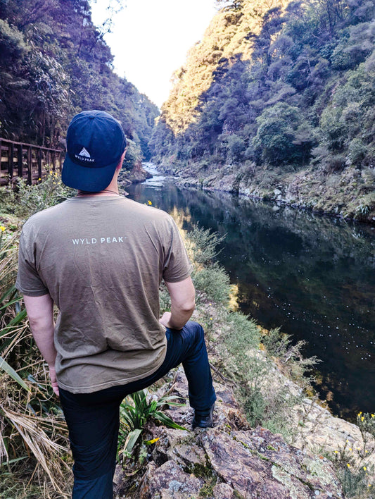 Alexander standing in a Wyld Peak tee, gazing out over the river at Karangahake Gorge, New Zealand, surrounded by rugged natural beauty