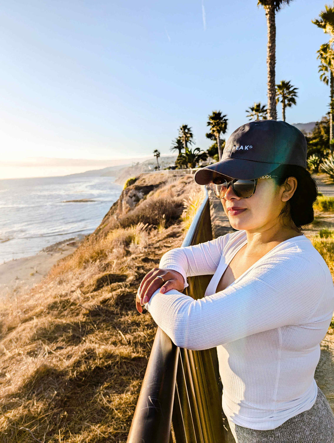 Mexican woman standing on the San Pedro cliffs in a Wyld Peak signature cap—adventuring with ocean views and coastal breeze
