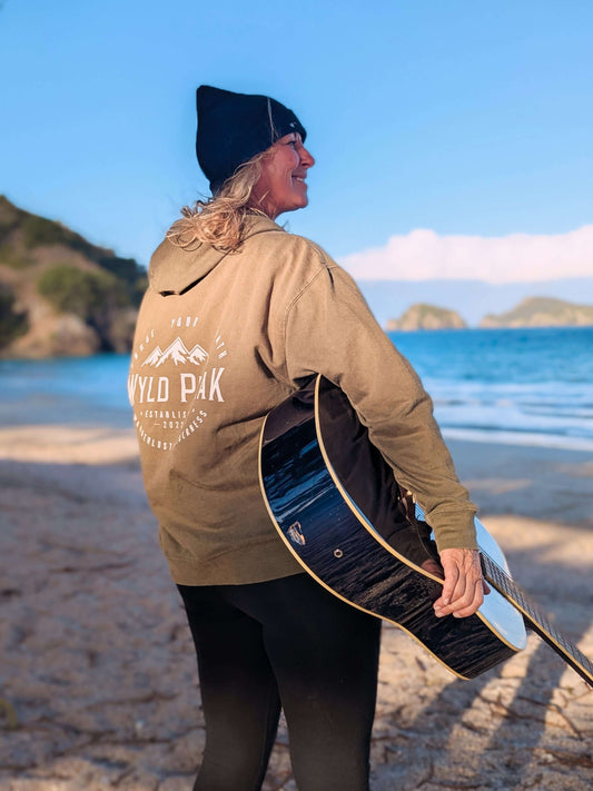 Sim Bastick wearing a Wyld Peak Organic Hoodie with a guitar, standing on the beachfront at Matauri Bay, New Zealand