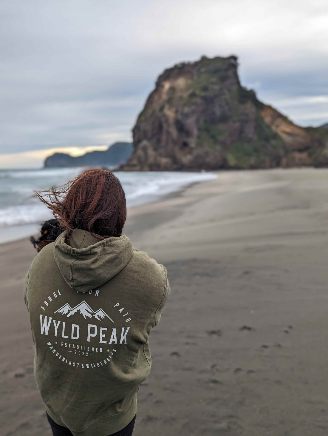 Yuzu wearing a Wyld Peak classic organic hoodie, gazing over the wild Piha coastline in New Zealand