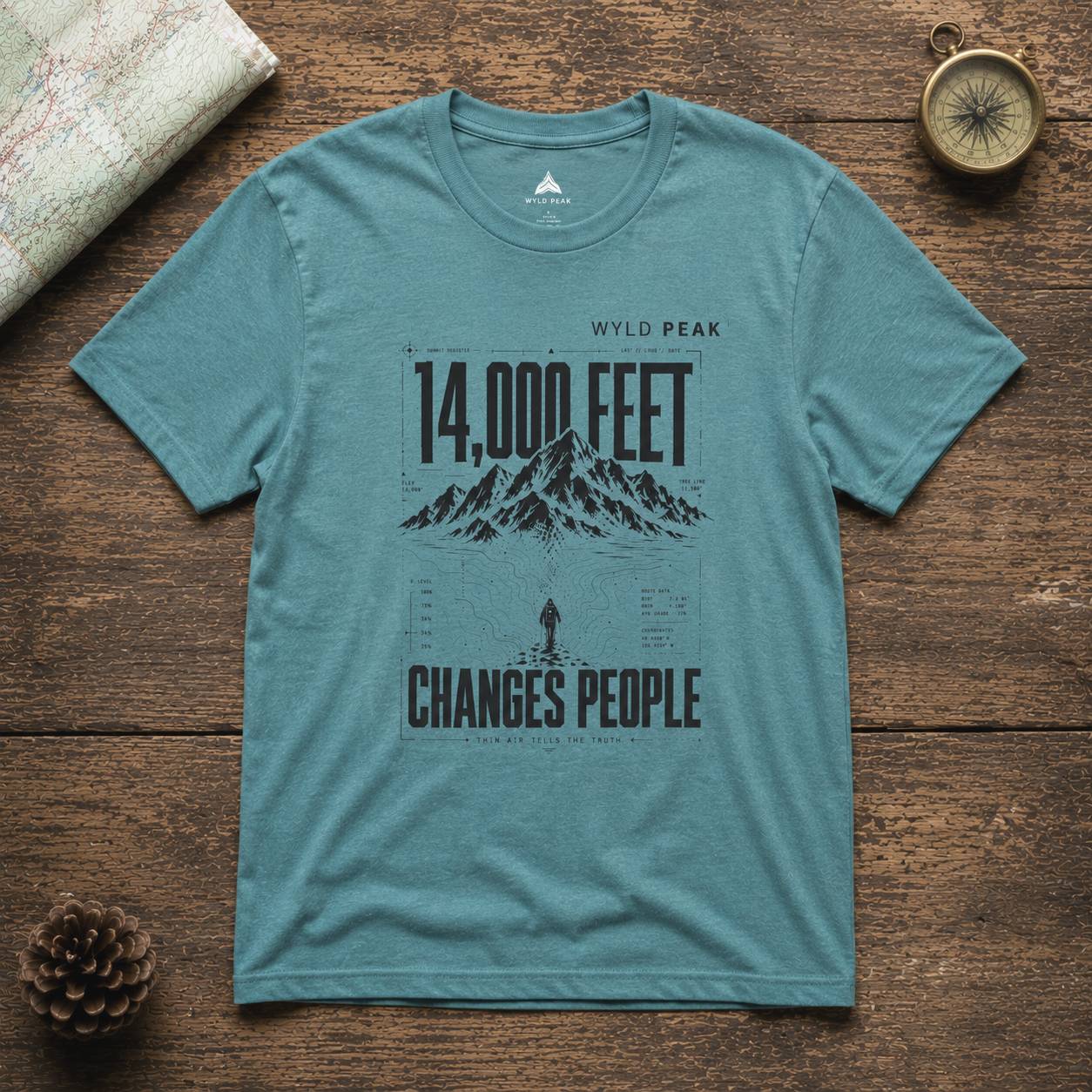 Teal t-shirt with mountain graphic and text on a wooden surface with map and compass.
