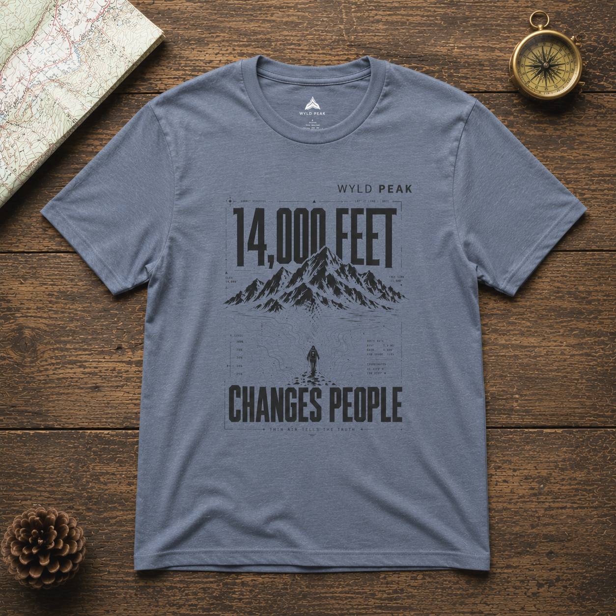 Gray t-shirt with mountain graphic and text on a wooden surface with map and compass.