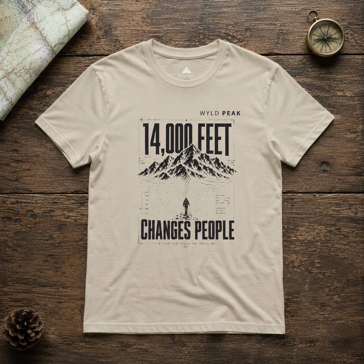 Beige t-shirt with mountain graphic and text on a wooden surface with map and compass.