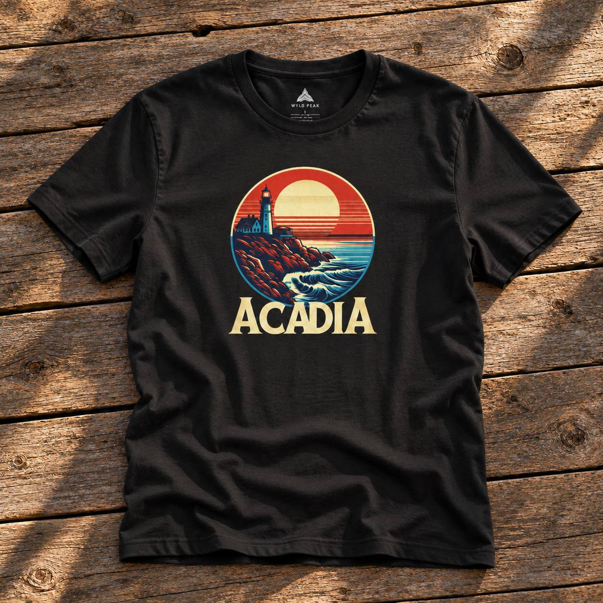 A black short-sleeve graphic t-shirt laid flat on a rustic wooden plank surface. The shirt features a circular retro-style print of a lighthouse on a rocky coast under a sunset, with the word 'ACADIA' printed below in a bold cream font.