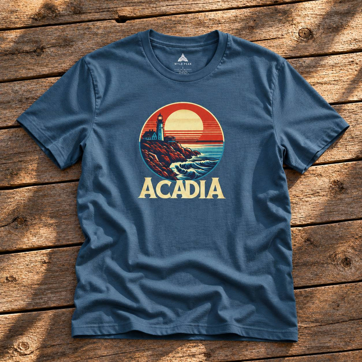 A slate blue short-sleeve t-shirt with a retro circular graphic of a lighthouse and sunset, featuring the word 'ACADIA' in yellow below it, laid flat on a rustic wooden plank background.