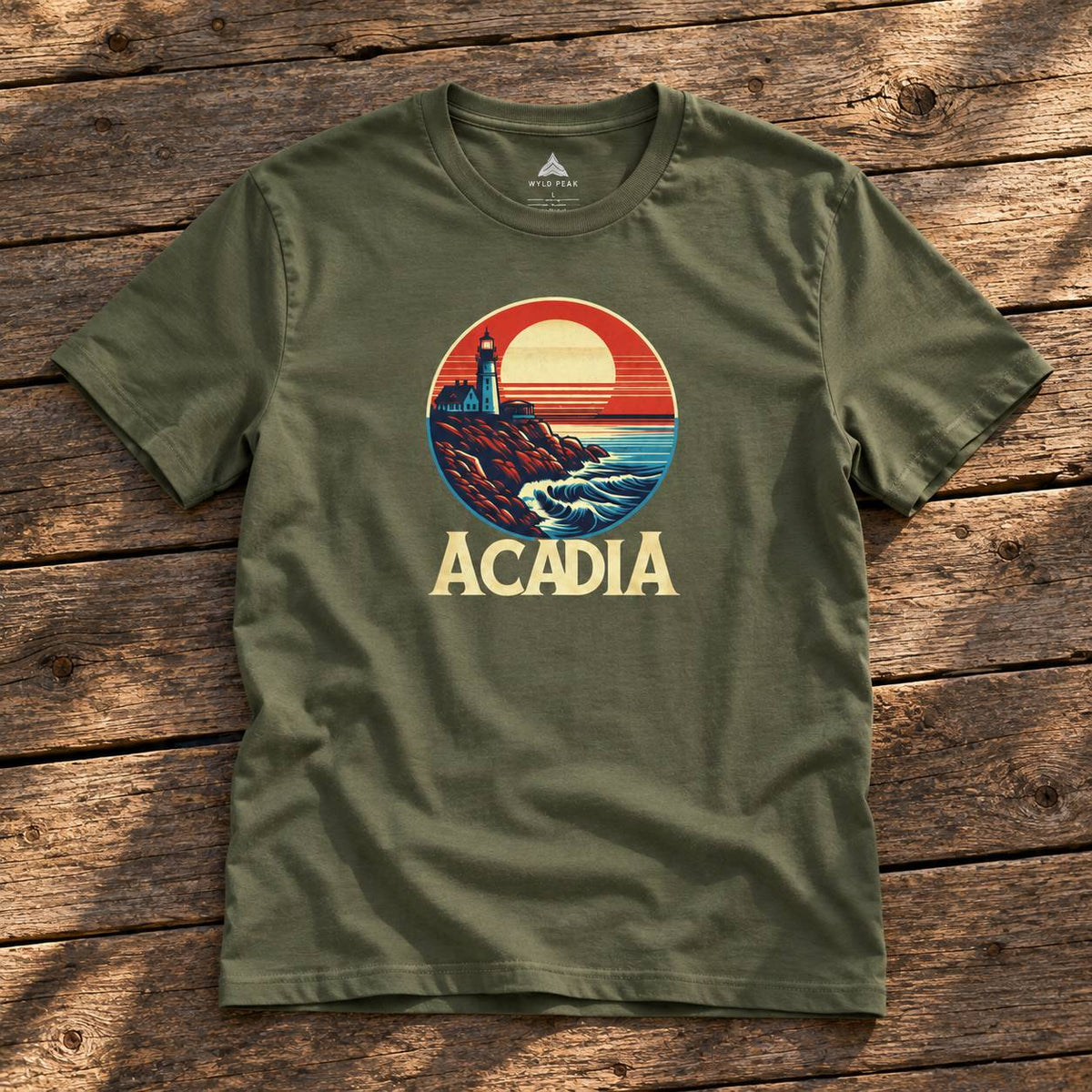 An olive green crewneck t-shirt with a retro circular graphic of a lighthouse on a rocky coast at sunset, with the word 'ACADIA' printed below in beige. The shirt is laid flat on a rustic wooden plank background.