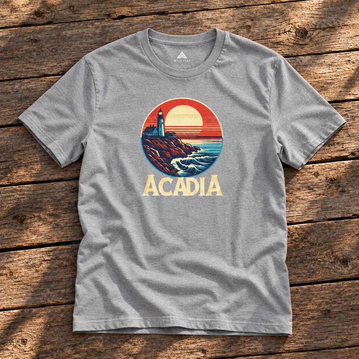 A heather gray crewneck t-shirt featuring a retro circular graphic of a lighthouse on a rocky coast under a sunset, with the word 'ACADIA' printed below in beige. The shirt is laid flat on a rustic wooden plank background.