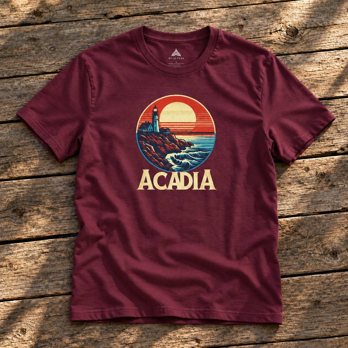 A maroon short-sleeve t-shirt with a retro circular graphic of a lighthouse and sunset, with the word 'ACADIA' printed below, laid flat on a rustic wooden plank surface.