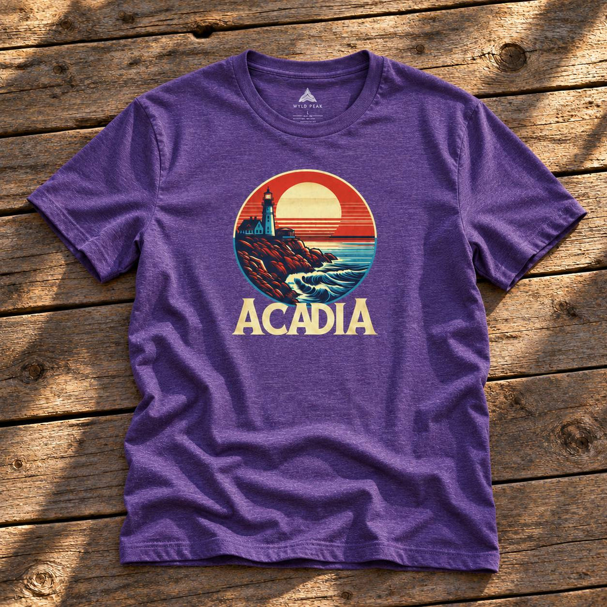 A purple heather crewneck t-shirt with a retro circular graphic of a lighthouse and sunset, with the word 'ACADIA' printed below, laid flat on a rustic wooden plank surface.