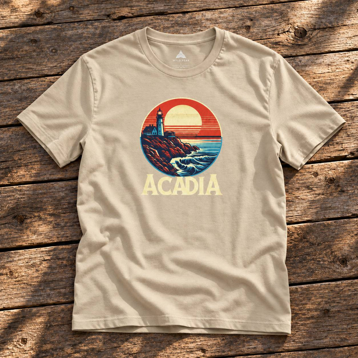 A beige short-sleeve t-shirt laid flat on a rustic wooden plank surface, featuring a circular retro-style graphic of a lighthouse and ocean sunset with the word 'ACADIA' printed below.