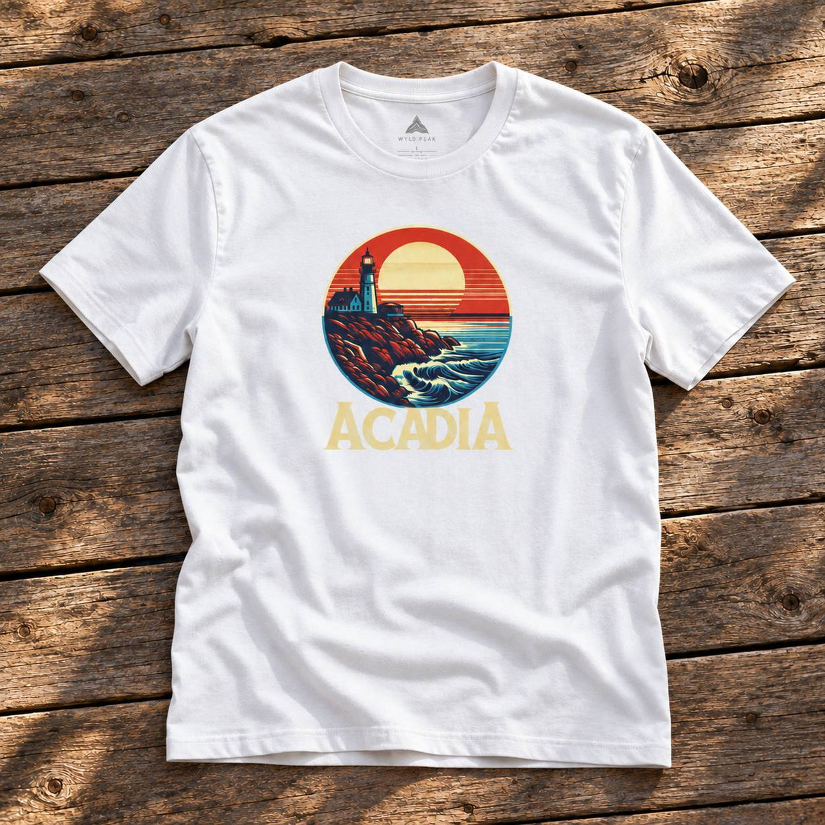 A white short-sleeve t-shirt with a retro-style circular graphic of a lighthouse and sunset, with the word 'ACADIA' printed below in yellow, laid flat on a rustic wooden plank background.