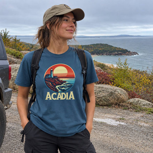 A woman wearing a slate blue t-shirt with a circular 'ACADIA' coastal graphic stands outdoors on a rocky path overlooking the ocean, with a car partially visible on the left.