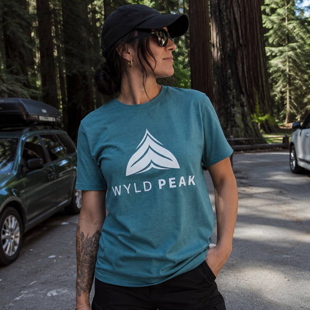 Person wearing a teal 'WYLD PEAK' t-shirt in a forest setting with vehicles in the background.