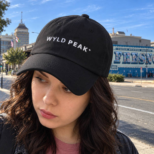 A woman wearing a black baseball cap with 'WYLD PEAK' embroidered in white on the front, photographed outdoors on a city street under a blue sky.