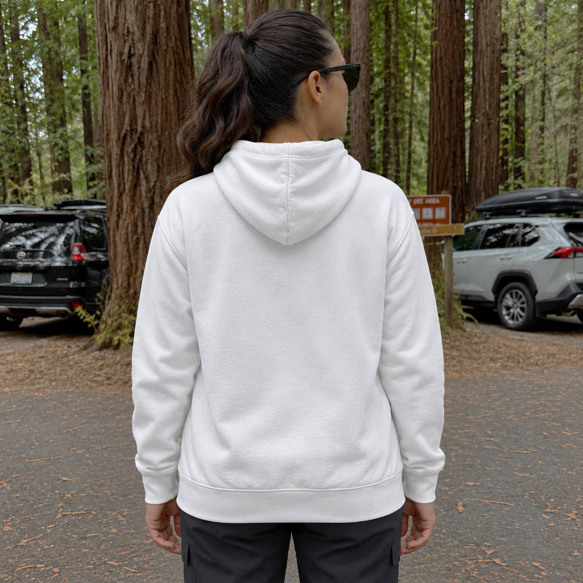 A woman viewed from behind wearing a plain white pullover hoodie in an outdoor forest parking lot with trees and parked cars in the background.
