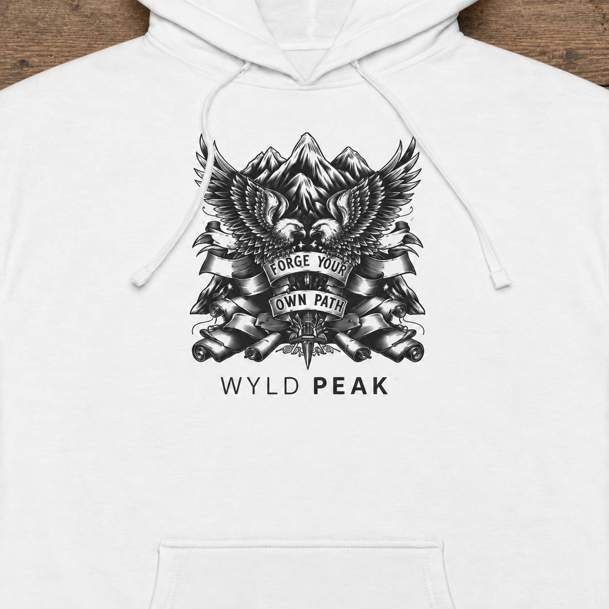 White pullover hoodie with a black graphic print featuring mountains, an eagle, banners reading 'Forge Your Own Path,' and the text 'WYLD PEAK' below, shown on a wooden background.