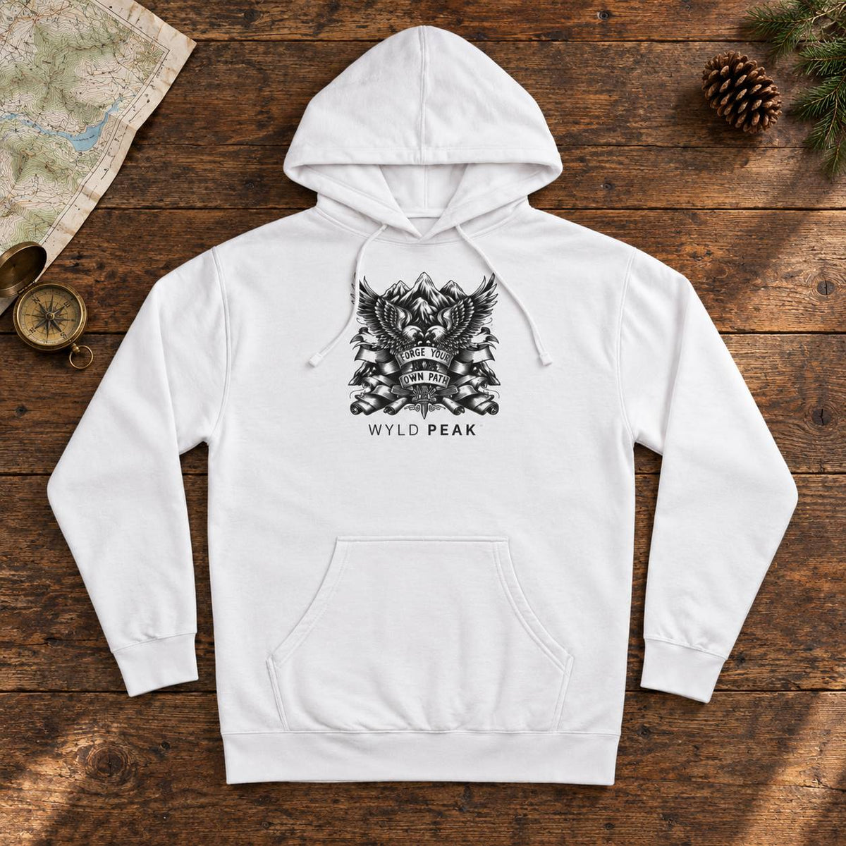 A white pullover hoodie with a black graphic print of an eagle and crossed rifles above the text 'WYLD PEAK', laid flat on a rustic wooden surface with a map, compass, pinecone, and evergreen branches around it.