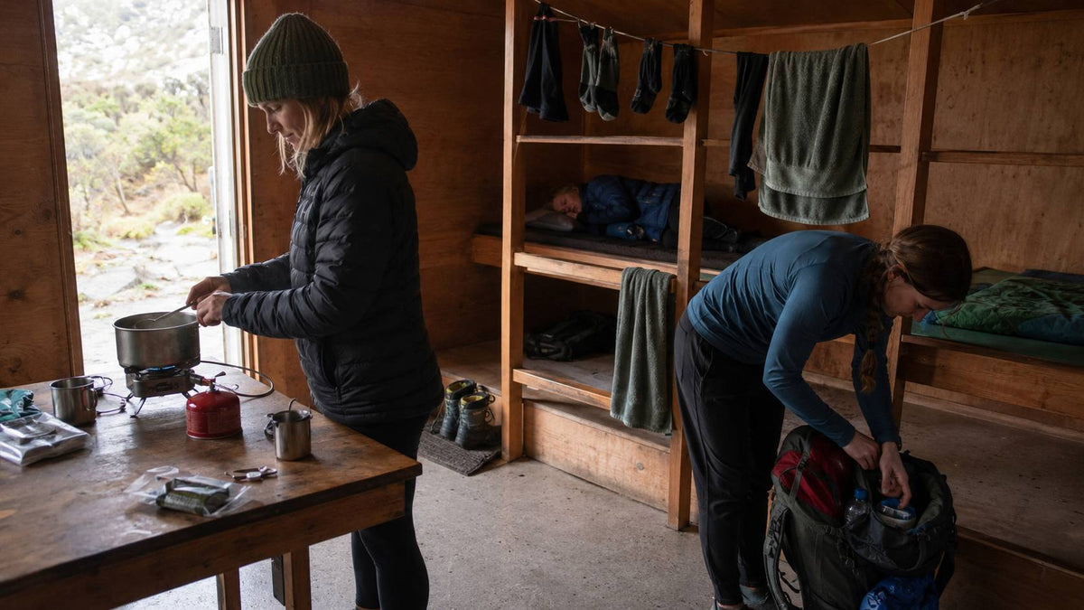 Two people preparing food and packing gear in a wooden cabin.