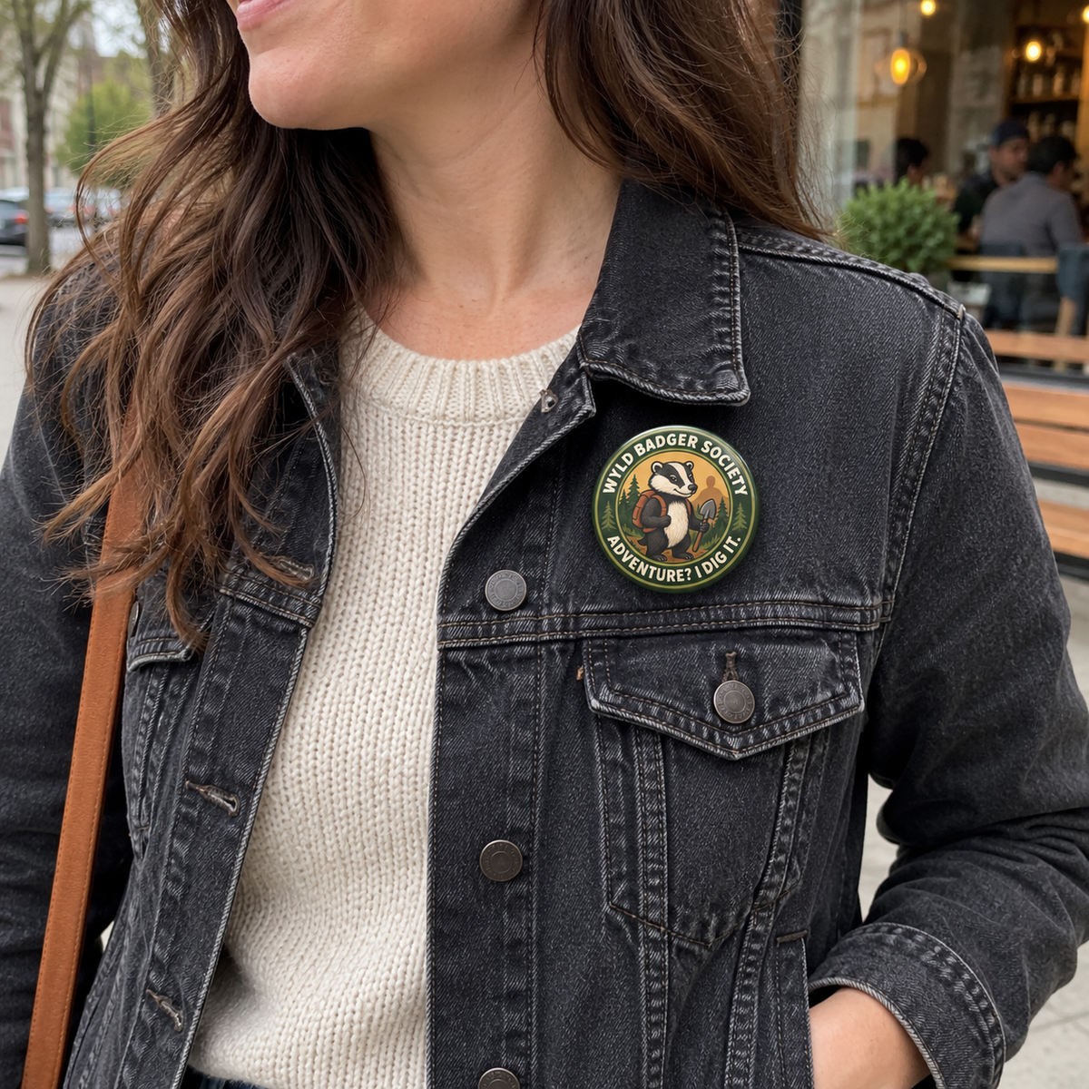 A close-up of a woman wearing a black denim jacket over a cream knit sweater, featuring a circular 'Wyld Badger Society' pin on the left chest, with an outdoor street background.
