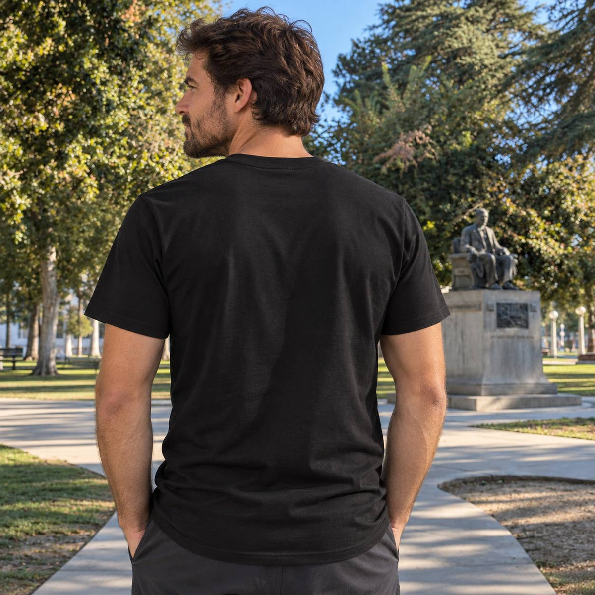 A man wearing a plain black short-sleeve t-shirt, viewed from behind, standing on a paved path in a sunny park with trees and a statue in the background.