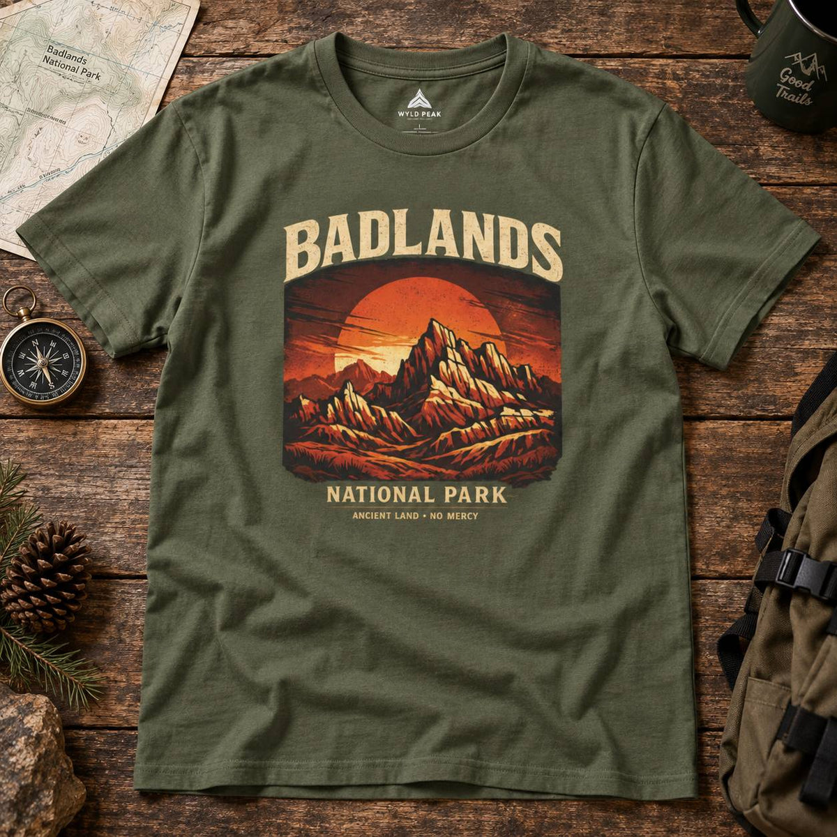 An olive green crewneck t-shirt featuring a retro-style graphic of the Badlands National Park sunset, styled in a flat-lay on a rustic wooden table with a map, compass, pinecone, and backpack.