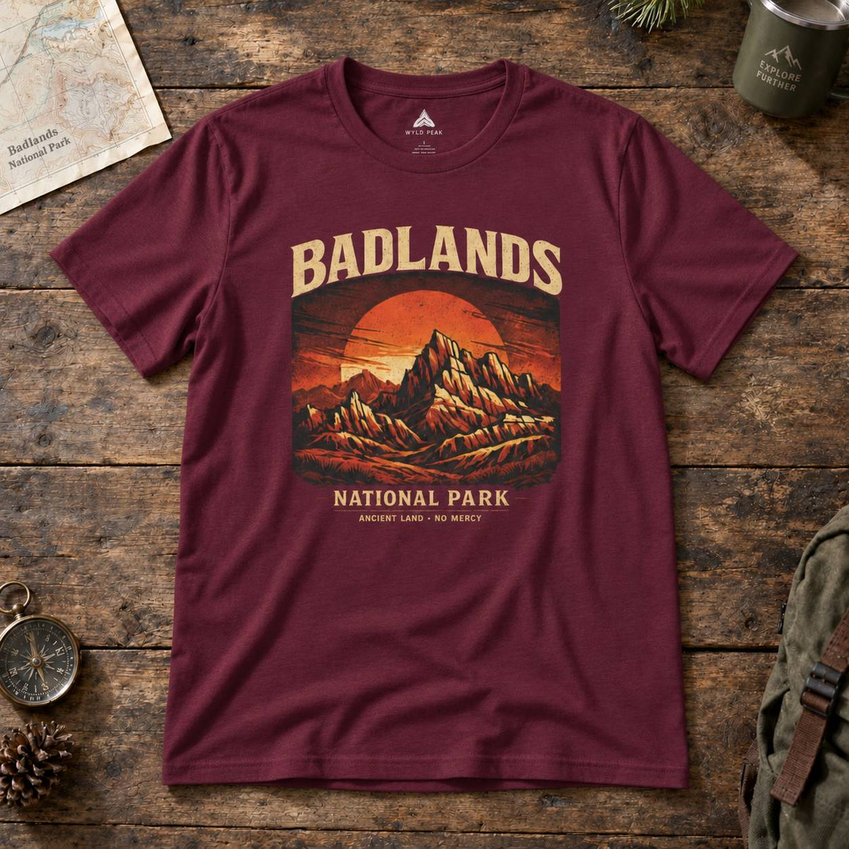 A maroon short-sleeve graphic t-shirt laid flat on a rustic wooden surface. The shirt features a retro-style mountain sunset print with the text 'BADLANDS NATIONAL PARK' and 'ARIZONA LAND OF THE MOUNTAINS'.