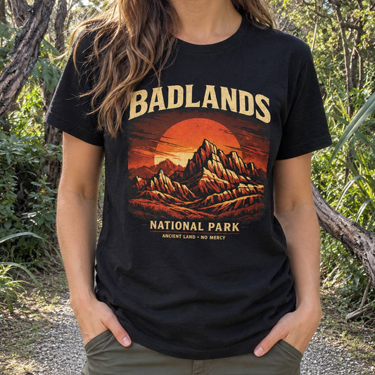 A woman wearing a black graphic t-shirt featuring a retro-style illustration of Badlands National Park with an orange sunset and beige text, set against a natural outdoor background of trees and foliage.